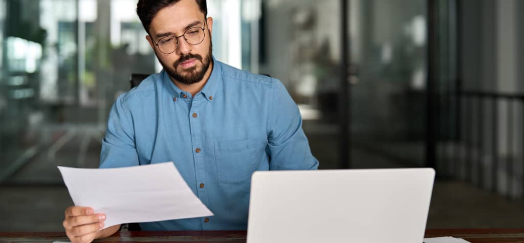 A person in a blue shirt sits at a desk holding documents, with a laptop open, in a modern office setting.