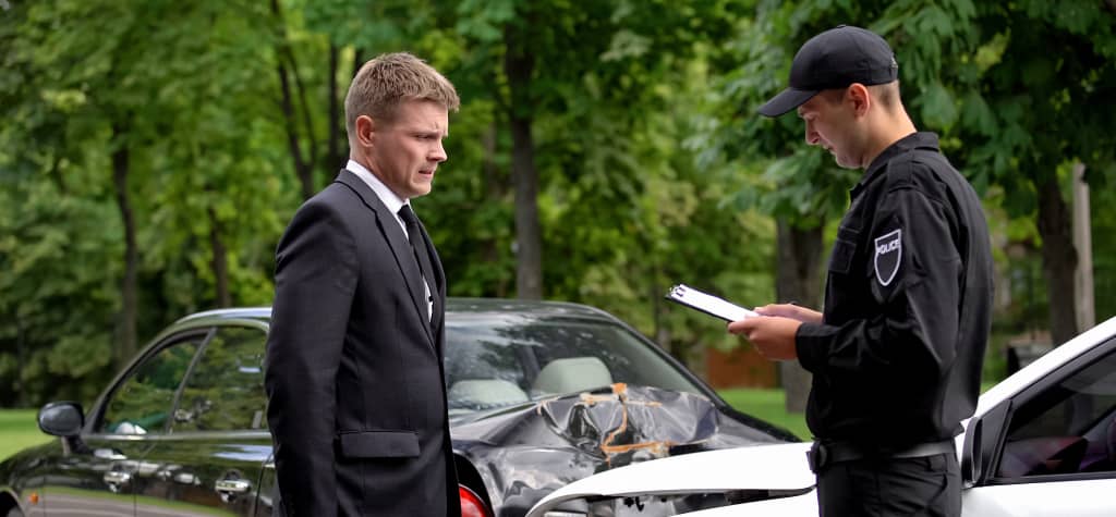 A police officer takes notes while discussing an auto accident with a man in a suit next to a damaged car in a green park setting.