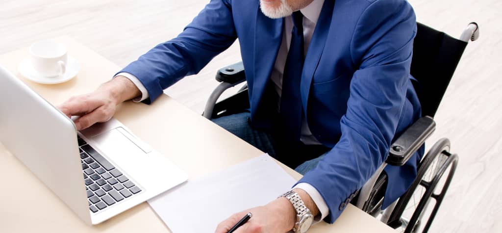 A man in a blue blazer, sitting in a wheelchair, works on a laptop while taking notes, with a cup of coffee nearby.