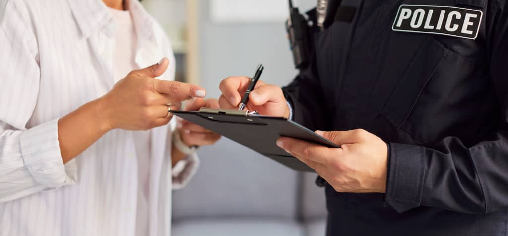 A police officer in uniform takes notes on a clipboard while a woman points, engaged in a discussion in an indoor setting.